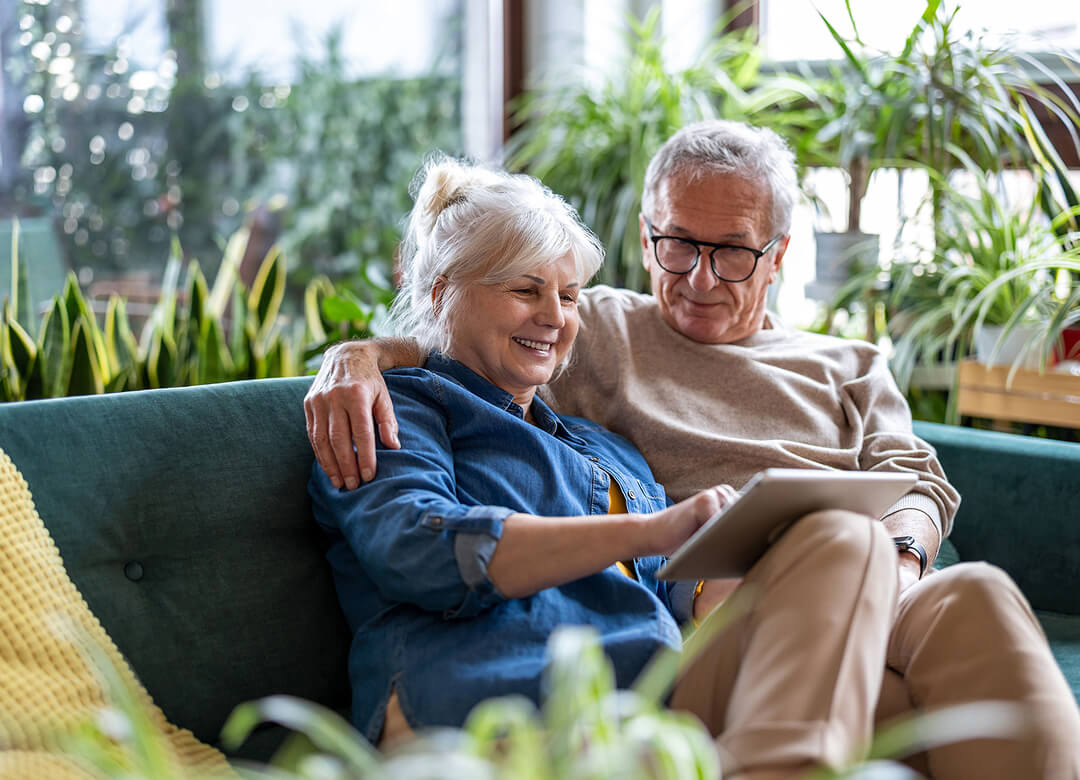 An older couple sits on a couch surrounded by plants, looking at a tablet together.