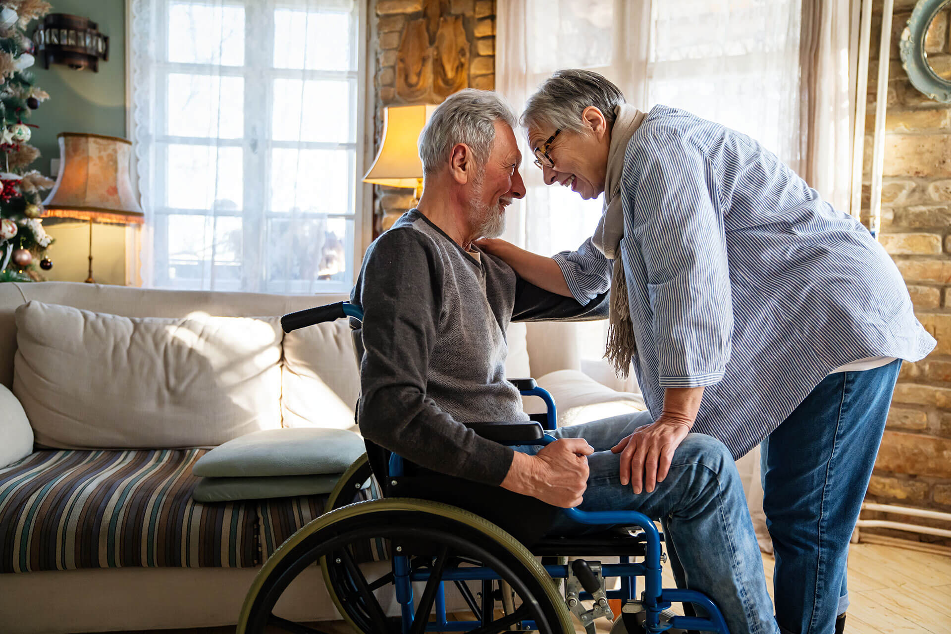 An elderly woman leans forward with a gentle smile, touching the shoulder of an elderly man sitting in a wheelchair. They are in a warmly lit living room with a couch, pillows, and a window in the background.