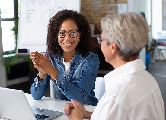 Two women are sitting at a table in a brightly lit office. One, wearing a denim shirt and glasses, smiles and clasps her hands. The other, with short gray hair, faces her and listens attentively. A whiteboard is visible in the background.